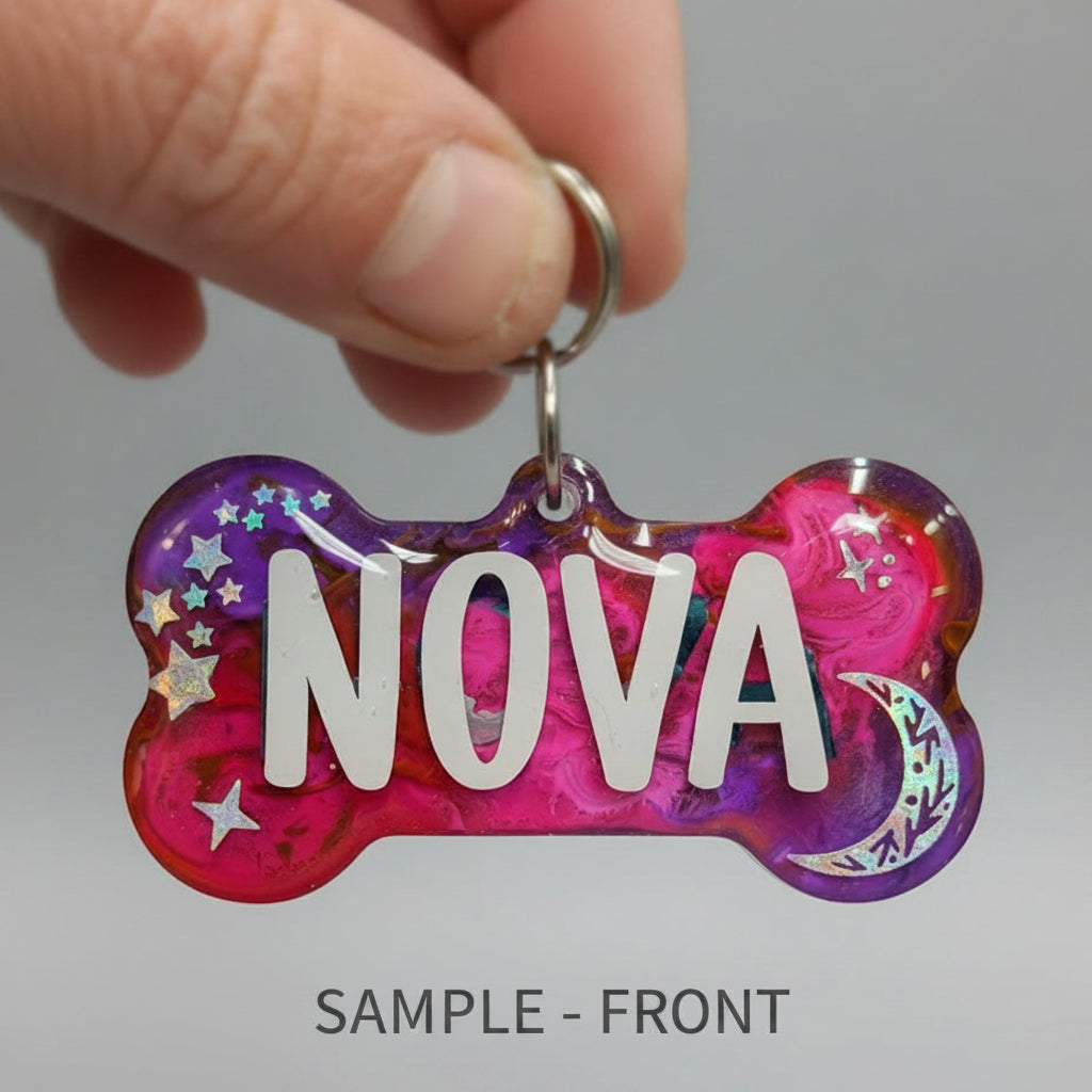 A hand holding a bone-shaped pet ID tag with the name 'NOVA' in white letters. The tag has a pink and purple glittery background with holographic star and crescent moon accents, attached to a silver ring, set against a solid purple background.