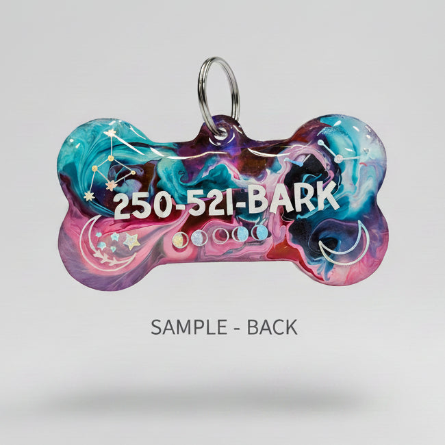 A hand holding a bone-shaped acrylic pet ID tag with a colorful marbled pattern of blue, pink, purple, and red, featuring the text '250-521-BARK' and a silver keyring.