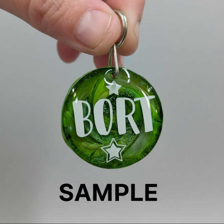 A close-up of a hand holding a round green bottle-cap-style keychain with the word “BORT” in white letters and white stars, against a purple background.